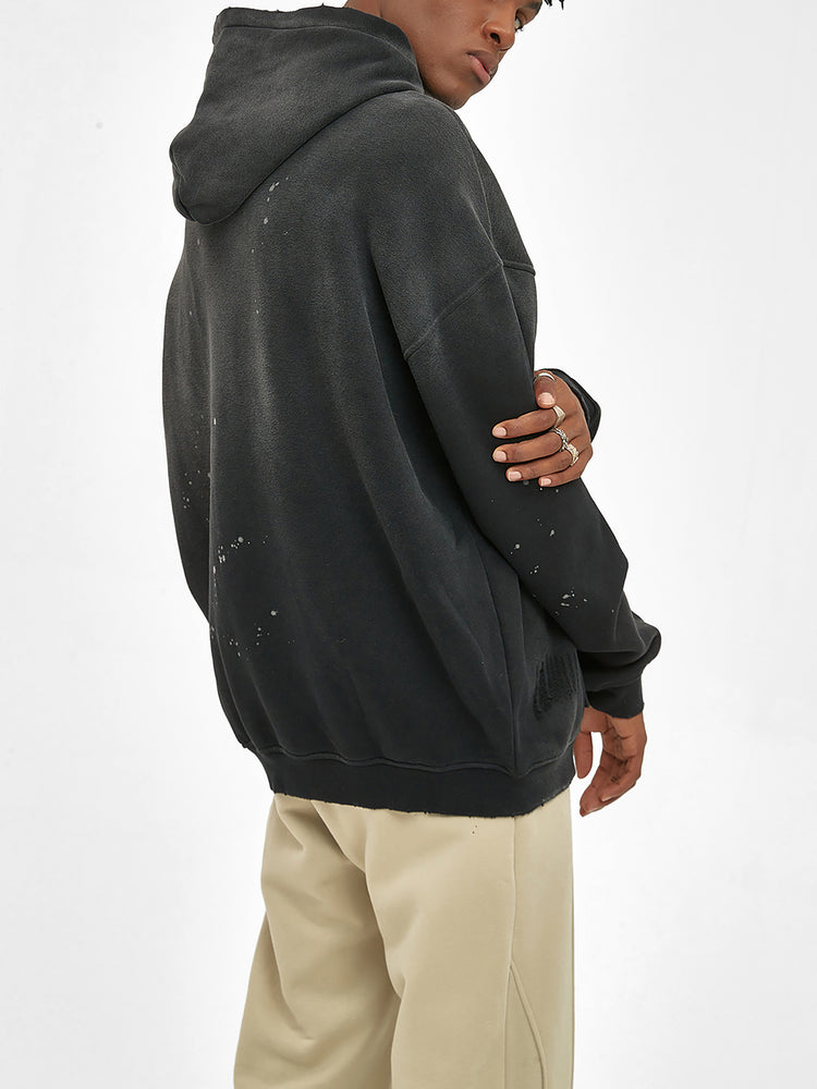 A person in the ODMPOD Baliveno Vintage Wash Frayed Fleece Hoodie (360 GSM) and beige pants stands turned away, glancing over their shoulder. The distressed black hoodie has small faded spots, set against a plain light background.