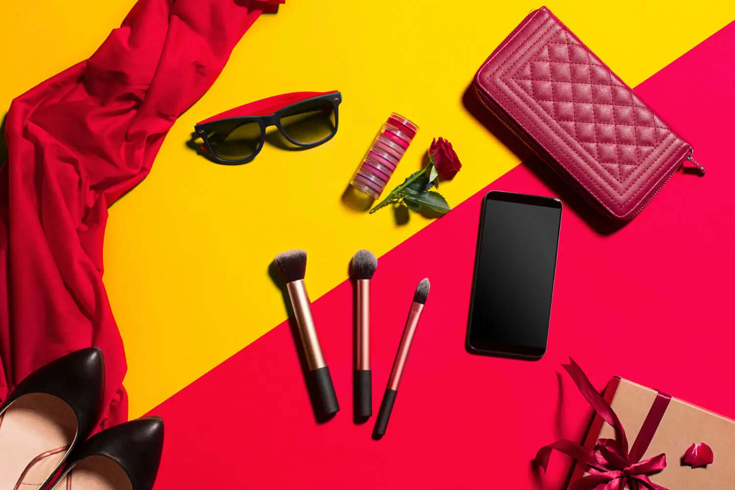 Fashion accessories and makeup essentials including a handbag, sunglasses, makeup brushes, and a red scarf laid out on a colorful background.