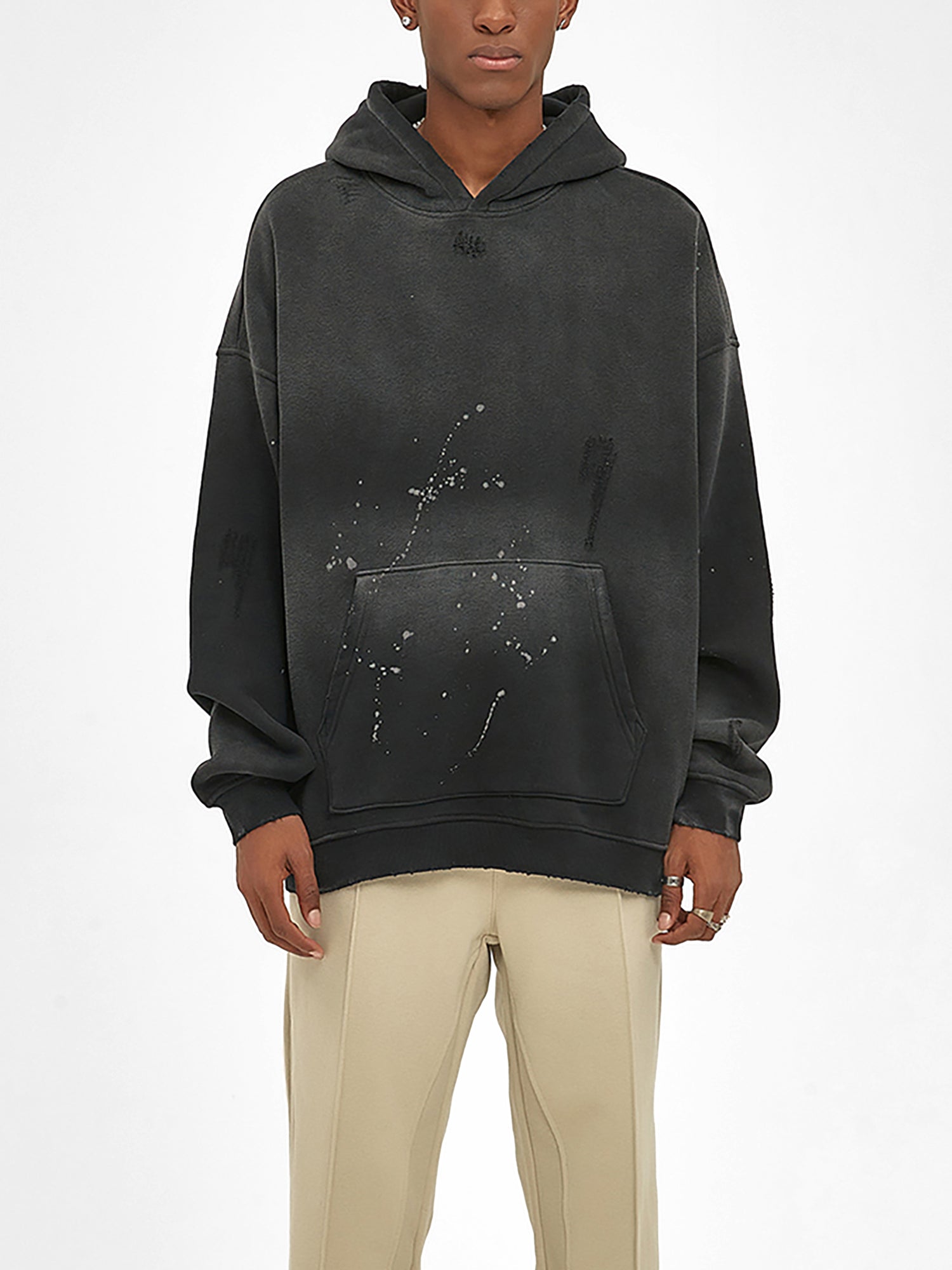 A person wears an oversized ODMPOD Baliveno Vintage Wash Frayed Fleece Hoodie (360 GSM) featuring a distressed, paint-splattered black design with beige pants, standing against a plain white background.