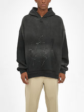 A person wears an oversized ODMPOD Baliveno Vintage Wash Frayed Fleece Hoodie (360 GSM) featuring a distressed, paint-splattered black design with beige pants, standing against a plain white background.