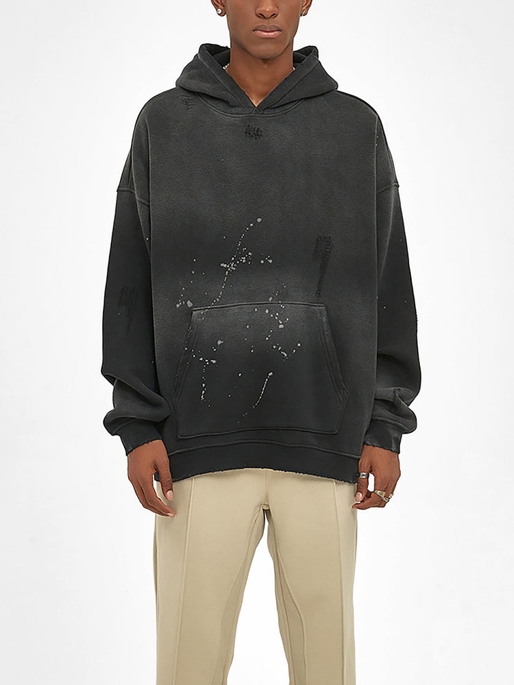 A person wears an oversized ODMPOD Baliveno Vintage Wash Frayed Fleece Hoodie (360 GSM) featuring a distressed, paint-splattered black design with beige pants, standing against a plain white background.