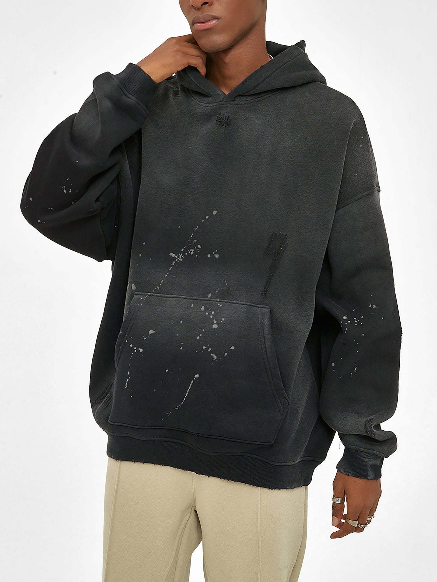 A person wears the oversized black ODMPOD Baliveno Vintage Wash Frayed Fleece Hoodie (360 GSM) with paint splatter details and beige pants, standing against a plain white background, hand touching face and head slightly tilted.