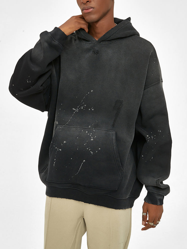 A person wears the oversized black ODMPOD Baliveno Vintage Wash Frayed Fleece Hoodie (360 GSM) with paint splatter details and beige pants, standing against a plain white background, hand touching face and head slightly tilted.