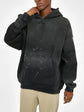 A person wears the oversized black ODMPOD Baliveno Vintage Wash Frayed Fleece Hoodie (360 GSM) with paint splatter details and beige pants, standing against a plain white background, hand touching face and head slightly tilted.