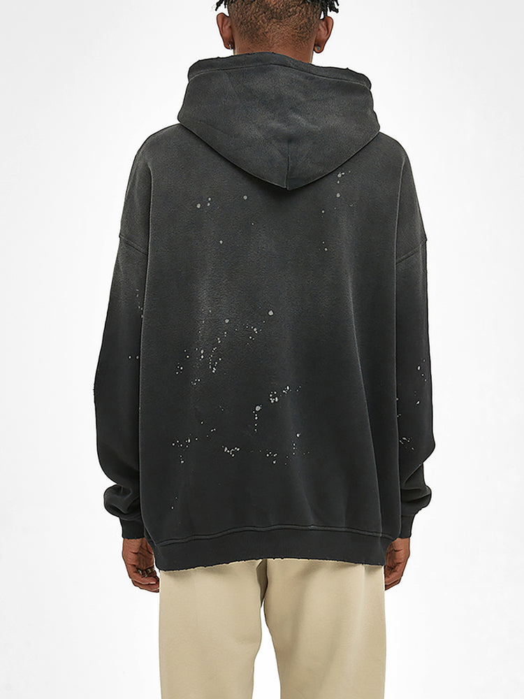 A person seen from behind, wearing the ODMPOD Baliveno Vintage Wash Frayed Fleece Hoodie (360 GSM) in black with white paint splatters on the back, paired with light beige pants against a plain white background.